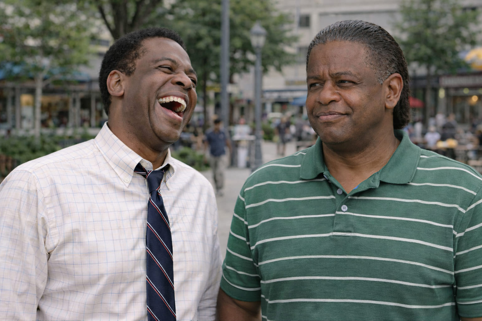 Two men outdoors sharing a broad laugh, one in black and one in a deep red shirt.